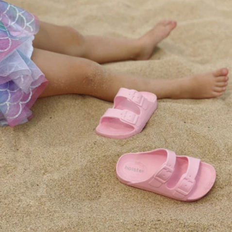 Pink sandals on a sandy surface with a child's legs wearing a colorful dress.